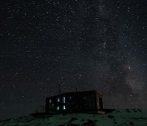 A large blue building, with some light showing through the windows sits on a snowy hilltop under a darky sky that is full of thousands of stars
