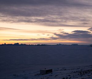 A small white building sits on the edge of a rocky, snow-covered landscape, with an expanse of sea ice stretching out to a group of icebergs on the horizon. The sky is filled with wispy clouds and the orange colours of sunset