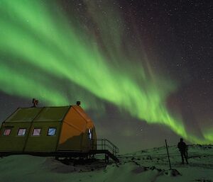A yellow, domed building is visible under a large green and purple aurora with the silhouette of a man standing beside it, looking away from the camera admiring the aurora.