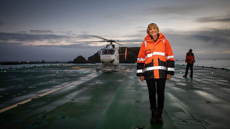 A woman in an orange high vis vest standing on a ship's deck with a helicopter behind.