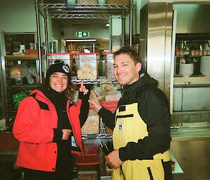 Two people standing in the staton kitchen pointing to some dried bananas on a shelf.