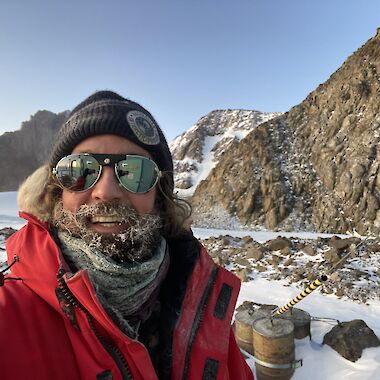 A man with ice in his beard, wearing a winter jacket and beanie, standing in front of snow-covered mountains.