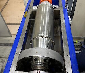 A long silver cylinder, covering an auger sits in a blue frame