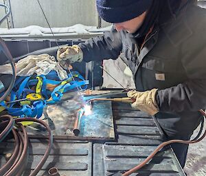 A man in a thick black jacket and blue beanie is using a blow torch to work with metal