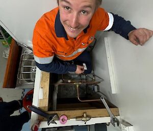 A man in orange high visibility clothing smiles up the camera from inside a crawl space