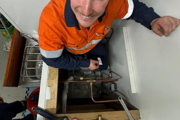 A man in orange high visibility clothing smiles up the camera from inside a crawl space