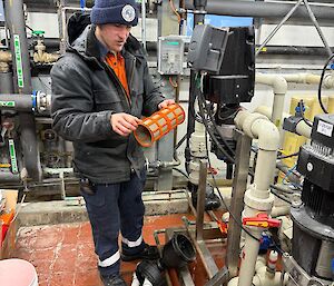 A man wearing orange high visibility clothing, a thick black jacket and a blue beanie is cleaning a small orange water filter