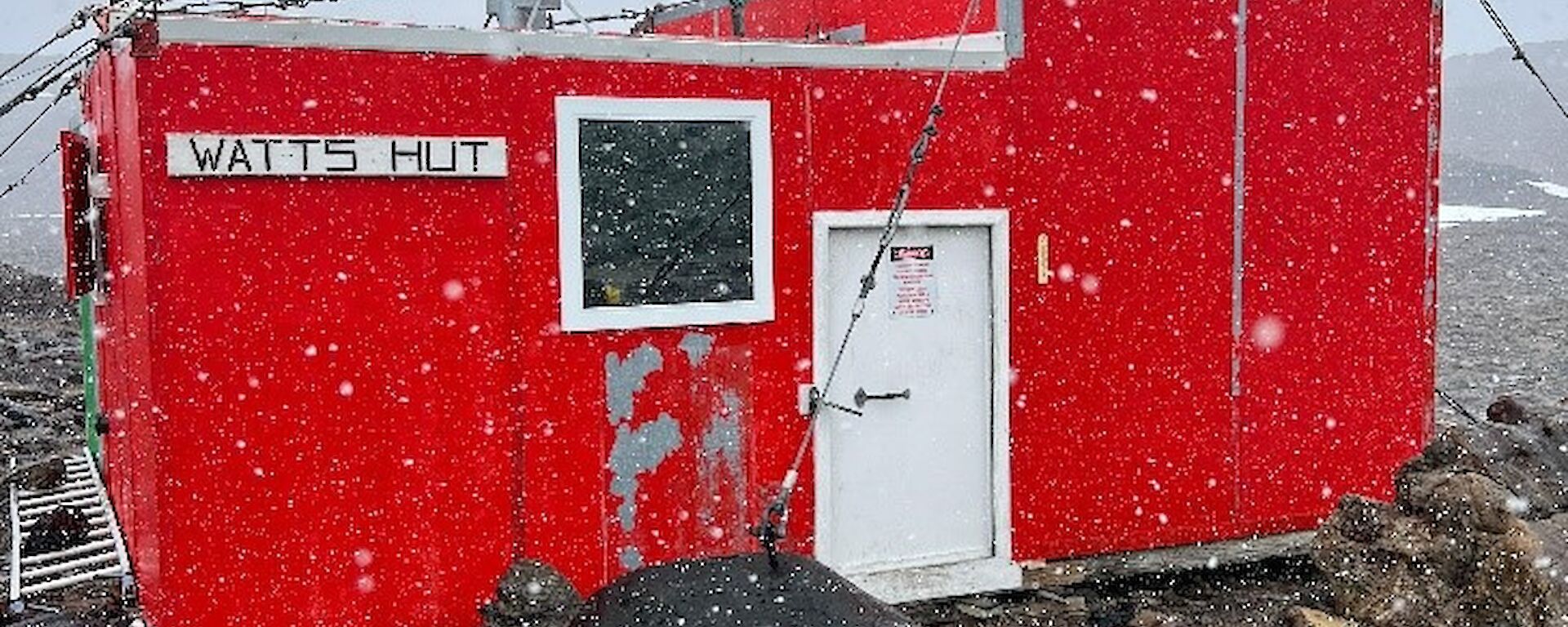 A square, red hut sits in a rocky landscape, snow is visible falling in the foreground