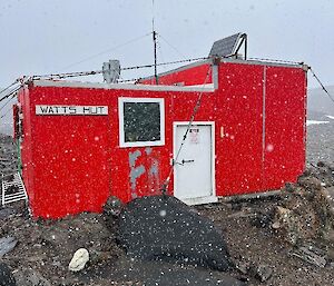 A square, red hut sits in a rocky landscape, snow is visible falling in the foreground