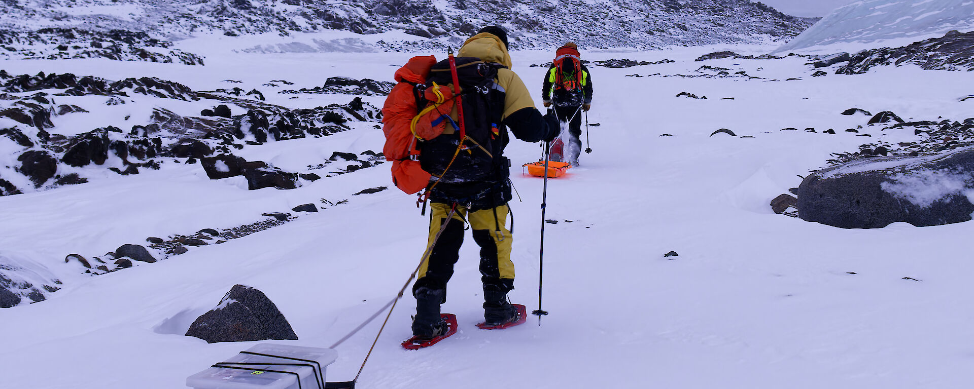 A man pulling a sled with a small plastic container on it through a snowy landscape.