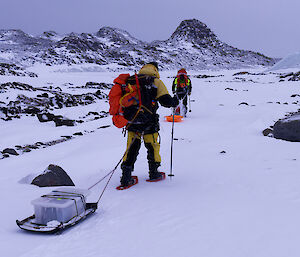 A man pulling a sled with a small plastic container on it through a snowy landscape.