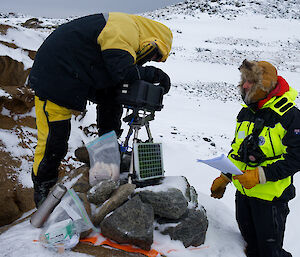 One man checks a camera on a tripod while another looks on with a paper manual in his hand.