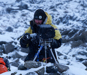 A man cleans snow off a camera, mounted on a tripod.
