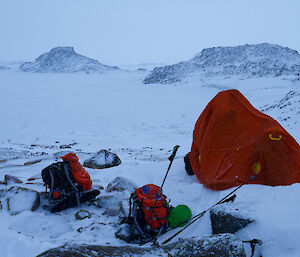 A red bag covering two people and a camera. Two packs sit outside in the snow.