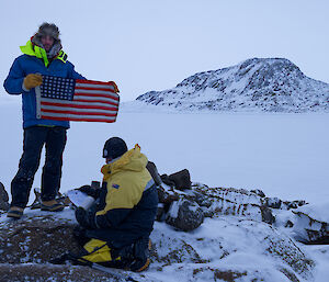 A man holding up a small American flag while another crouches on the ground beside a stone cairn.