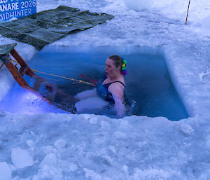 A woman is swimming in a square hole cut into sea ice, with a ladder to get in and out.