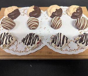 A wooden platter with paper doilies presents 12 petit four sweets.
