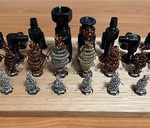 Two sets of chess pieces are set into a wooden board. One set is made from nuts and bolts and painted black, the other set is made from wire twisted into the shape of penguins