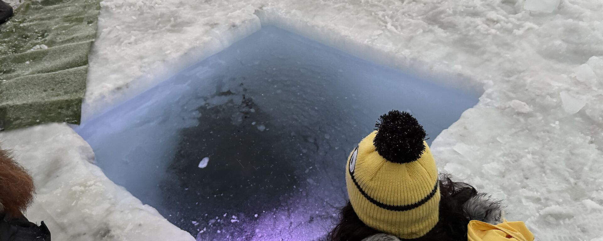 A person wearing a yellow jacket and beanie squats next to a hole cut into the sea ice.