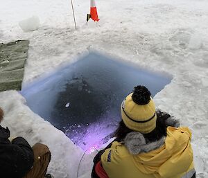 A person wearing a yellow jacket and beanie squats next to a hole cut into the sea ice.