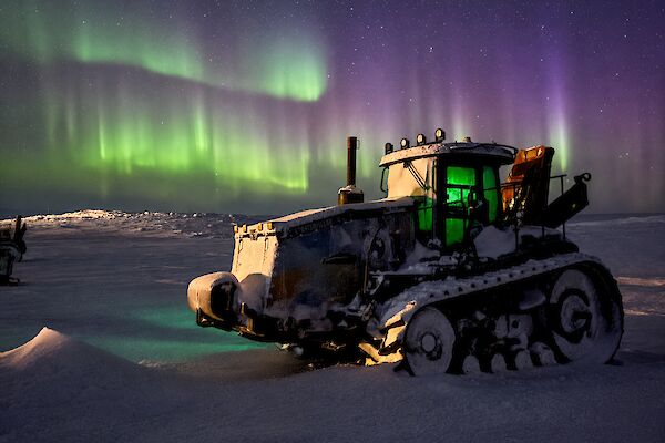 A snow-covered tractor with a green and purple aurora in the sky.