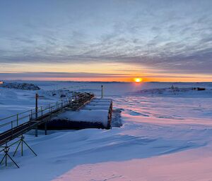 A rising sun on the horizon, with snow covered fuel drums in the foreground.