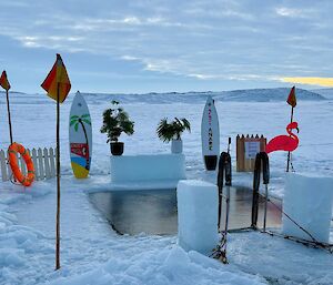 A square hole carved in the ice, surrounded by surfboards, a flamingo, flags and a life ring. A set of steps is positioned on the side for people to enter the hole.