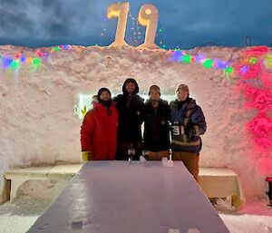 Four people posing in front of a wall build from snow blocks. The number 79 is on the top of the wall. Colourful lights are strung along the top of the wall.