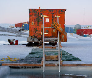 A red 'RMIT' van parked on the sea ice, behind Mawson's mid-winter swim hole that has been cut out of the sea ice.