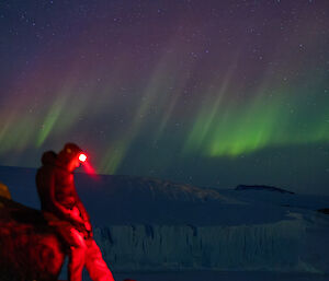 An expeditioner with a head torch shining a red light on the ground in the foreground, with an aurora and stars featuring in the night sky.