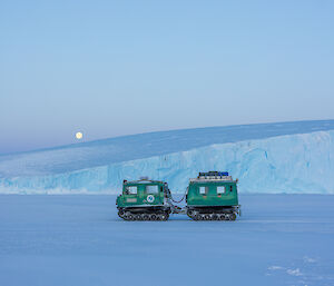 A green Hagglands over-snow vehicle sitting on the sea ice, with ice cliffs and full moon in the background.