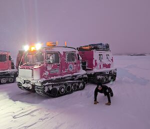 A person wearing a black jumper and beanie smiles up at the camera whilst doing a push up next to two pink and purple Hagglungs vehicles