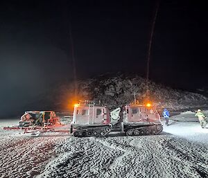 A pink vehicles towing a sled, both of which are covered in snow is parked on stippled snowy ice in front of a rocky hill. People are walking around on the ice in front of the vehicle