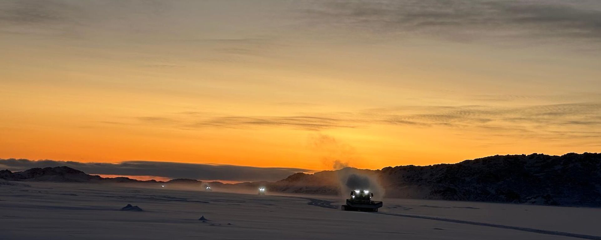 A wide picture of a flat expanse of snow, with a rocky island visible and a convoy of vehicles travelling towards the camera. The first vehicle is a snow groomer with its headlights on and a steamy fog coming from the exhaust. The other three vehicles are visible only as headlights behind them.
