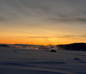A wide picture of a flat expanse of snow, with a rocky island visible and a convoy of vehicles travelling towards the camera. The first vehicle is a snow groomer with its headlights on and a steamy fog coming from the exhaust. The other three vehicles are visible only as headlights behind them.