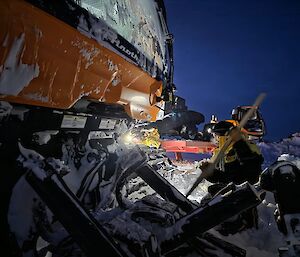 A person in a thick yellow and black beanie wearing a helmet tourch crouches with a shovel next to a large yellow vehicle that is partially covered in snow.
