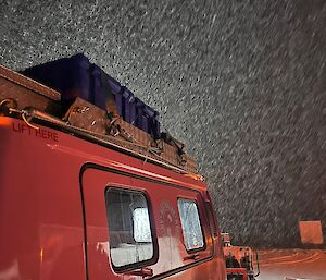 A pink vehicle is parked with its headlights lighting up the falling snow in front of it.