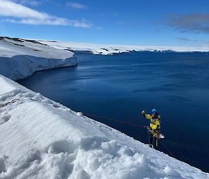 A man attached to a rope, part way down an ice cliff, with a blue bay below.
