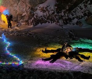 Colourful fairy lights decorating snow and rocks. A group of people are lying on the ground in a star shape.