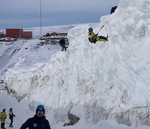 Two people abseiling down an ice cliff. Three people on the ground below.