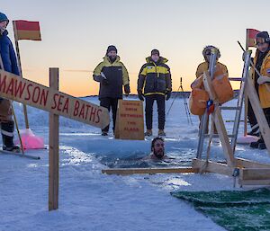 An expeditioner swimming in the sea ice hole, surrounded by warmly-dressed expeditioners standing around the hole