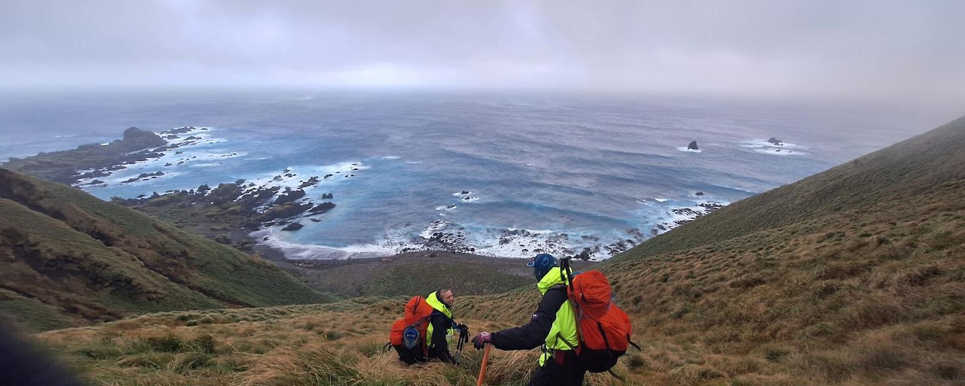 Two people help each other up a steep grassy slope with the wild sea behind them