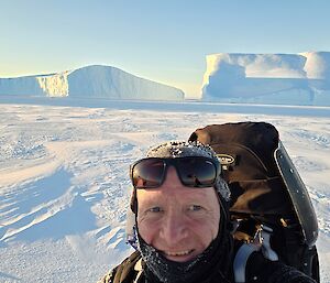 A man wearing a backpack with a pair of sunglasses pushed up onto his head is standing in a snowy landscape with large icebergs visible in the background. It is a bright and sunny day with a pale blue sky.
