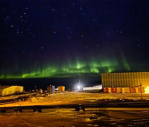 A dark blue sky, studded with stars is lit up by ribbons of green aurora hovering just above square buildings sitting in a snowy landscape.
