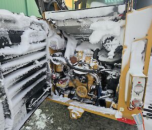 The engoine of a yellow plant vehicle is covered in snow