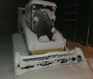 A yellow snow groomer is partially buried by snow. It is dark.