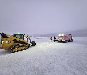 A pink Hagglunds vehicle and a small yellow skidsteer with a large auger drill attachment sit in a snowy landscape.