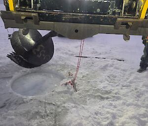 A large auger drill attachment is connected to the front of a small yellow bulldozer and starting to dig a hole, just over 1m wide in the sea ice