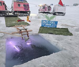 A square swimming hole has been cut out in the sea ice, with a ladder entering the water that is lit up. In the background are beach style decorations and two Hagglunds Vehicles