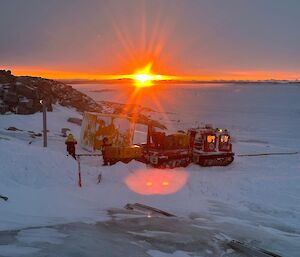 Two snow tractors stand on the snow with the sun rising behind them